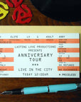 Ticket-style invitation for an 'Anniversary Tour' with decorative elements on a wooden surface.