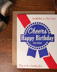 Birthday card with 'Cheers Happy Birthday To You' design on a wooden surface with money.