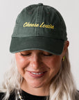 Person wearing a green cap with 'Cheese Louise' embroidered on it against a plain background