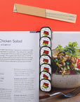 Sushi-themed bookmarks on a recipe book with a bowl of salad and fork on a red background