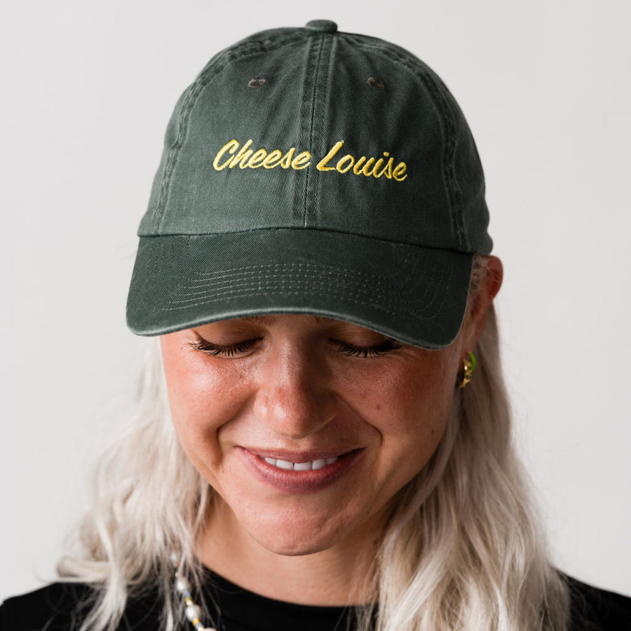 Person wearing a green cap with 'Cheese Louise' embroidered on it against a plain background