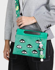Green handbag with eye designs and a black strap held by a person wearing a checkered jacket.
