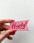 Hand holding a pink embroidered patch with 'Howdy!' text on a white background