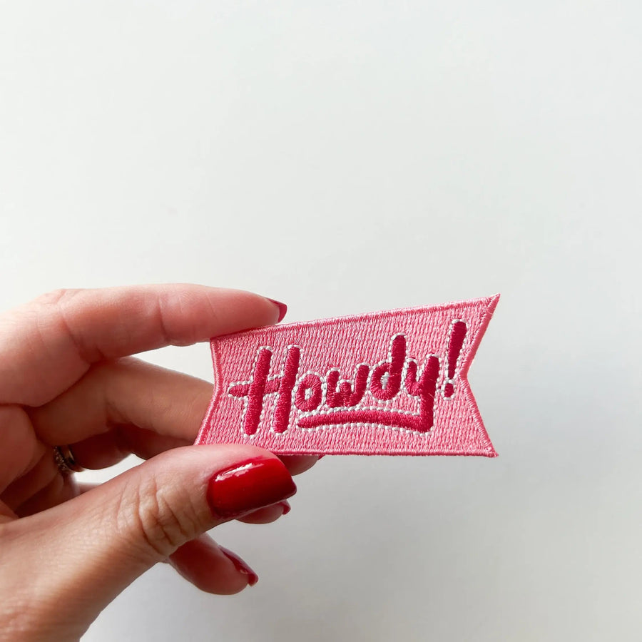 Hand holding a pink embroidered patch with 'Howdy!' text on a white background