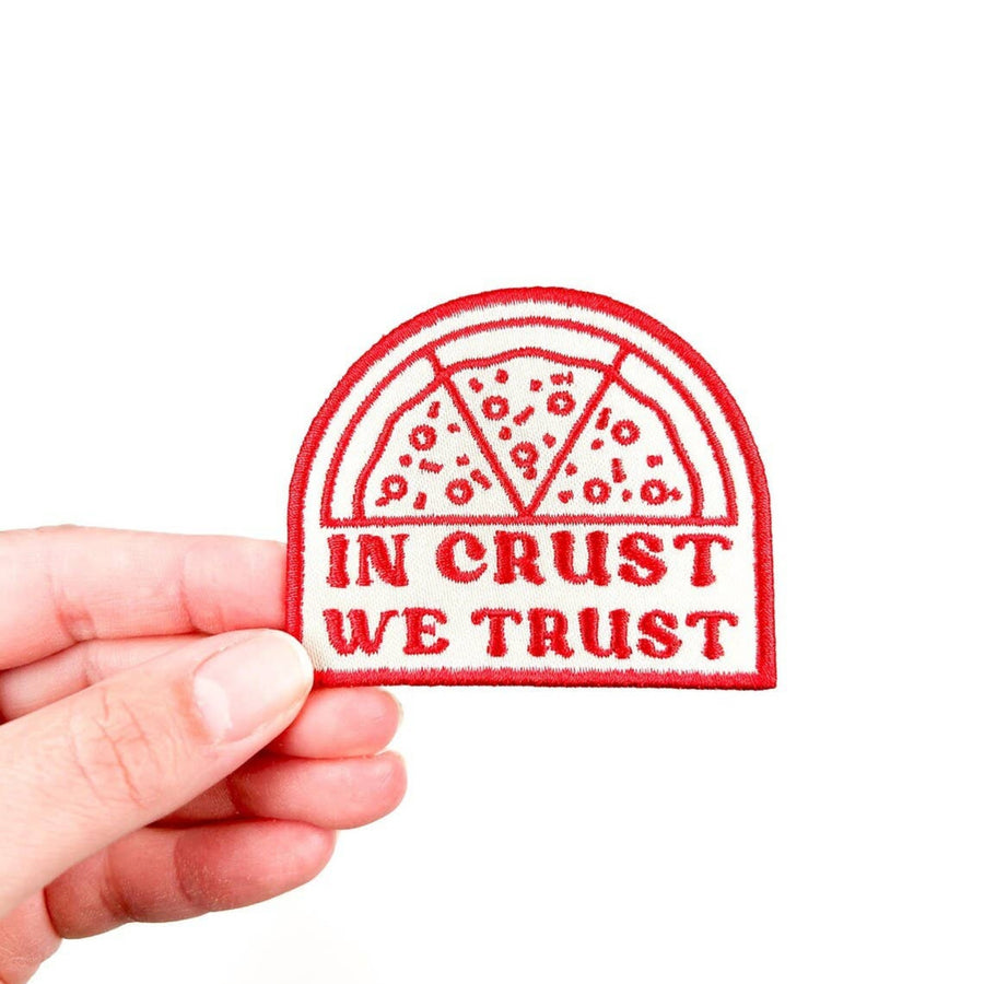 Patch with 'In Crust We Trust' text held by a hand on a white background