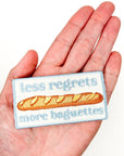 Embroidered patch with a baguette and text 'less regrets more baguettes' held in a hand.