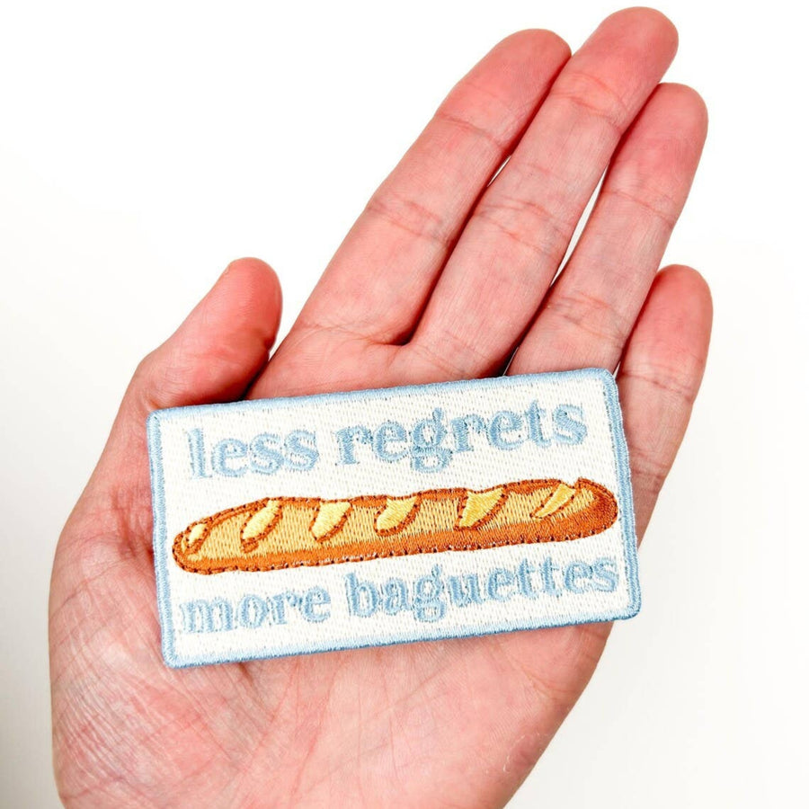 Embroidered patch with a baguette and text 'less regrets more baguettes' held in a hand.