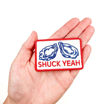 Patch with oyster design and 'SHUCK YEAH' text held in a hand on a white background