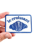 Patch with 'le croissant' text and croissant illustration held by a hand on a white background