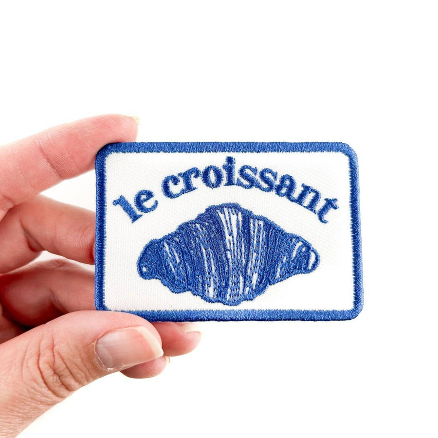 Patch with 'le croissant' text and croissant illustration held by a hand on a white background