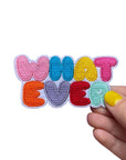 Colorful patch with 'WHAT EVER' text held by a hand on a white background