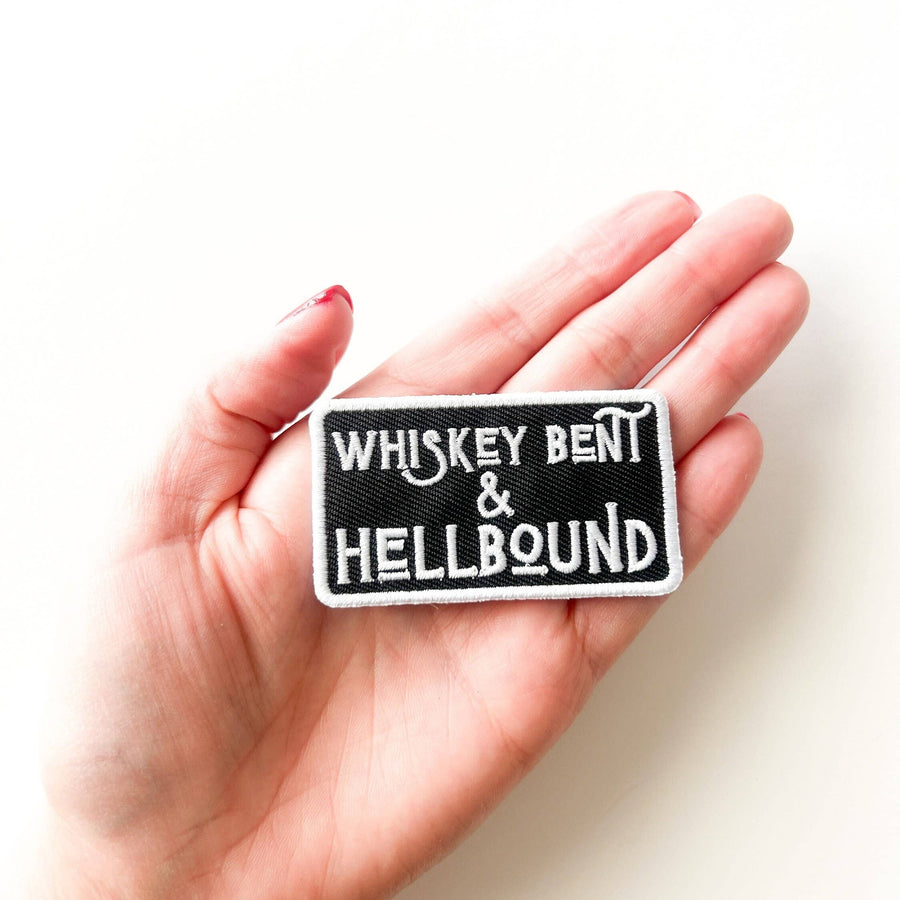 Hand holding a patch with 'Whiskey Bent & Hellbound' text against a white background